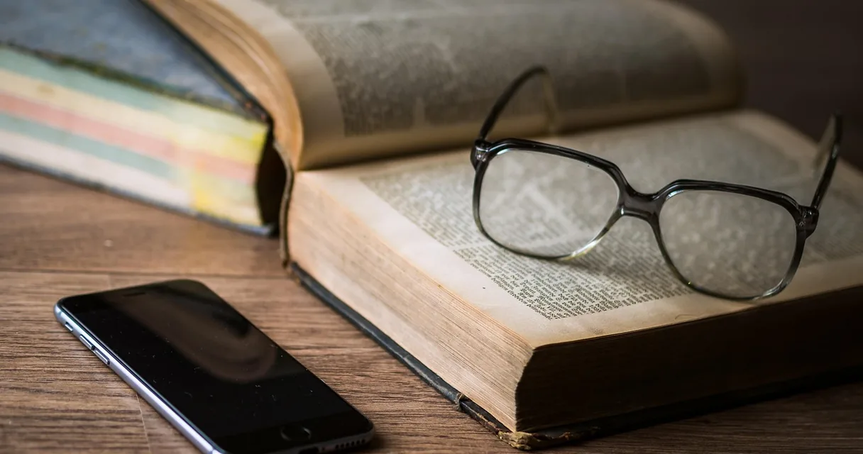 Livro aberto com óculos e um celular sobre uma mesa de madeira, ilustrando como checar notícias sobre tecnologia